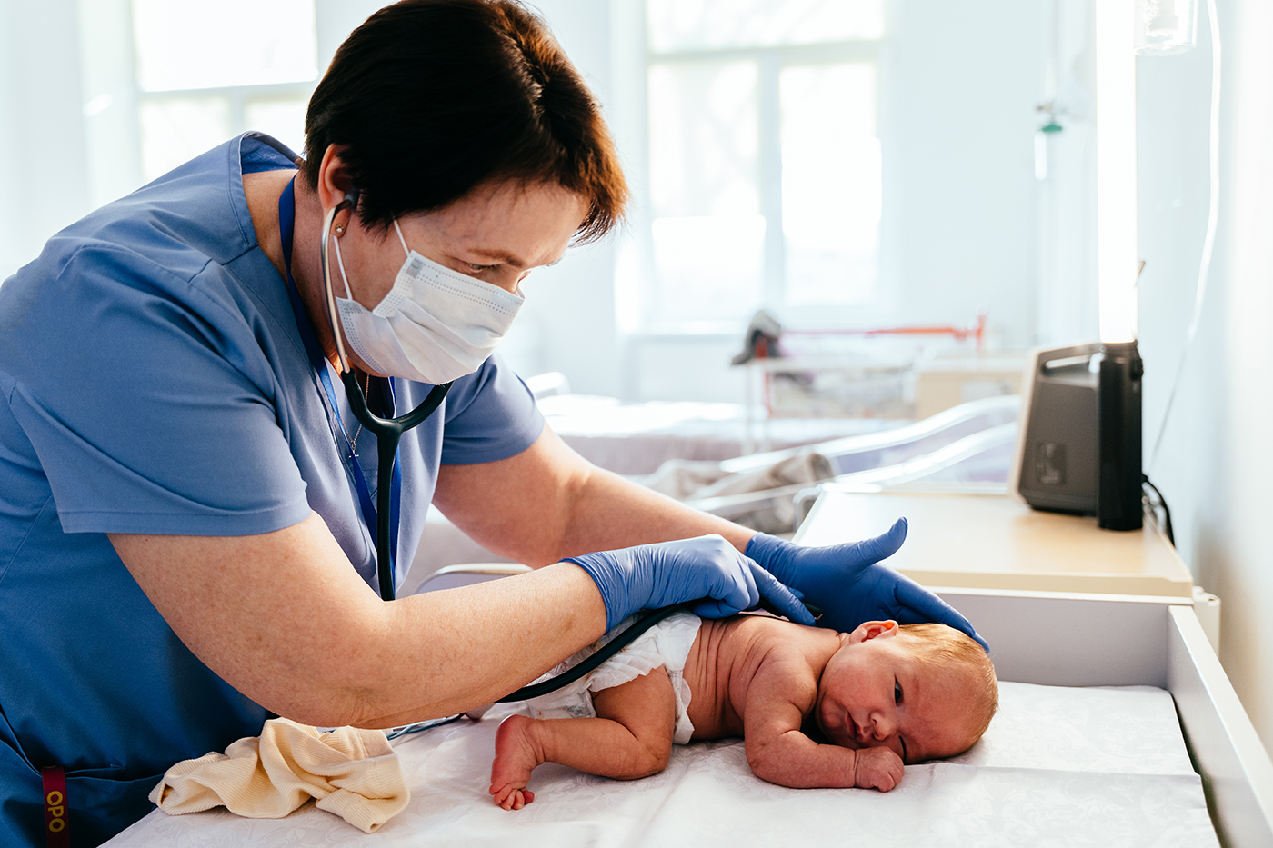 A healthcare worker in blue scrubs and gloves examines a newborn baby on a changing table in a medical setting. The worker uses a stethoscope while the baby lies on its stomach. True Joy Birthing