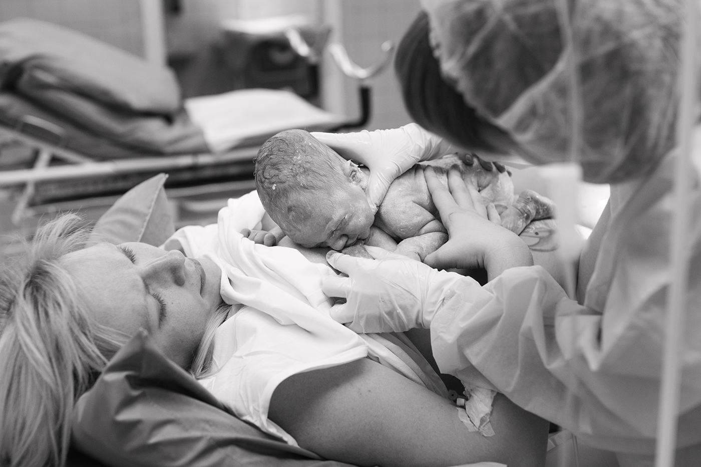 A newborn baby is placed on a woman’s chest immediately after birth, as a medical professional assists. Both adults wear hospital gowns, and the scene takes place in a medical setting. True Joy Birthing